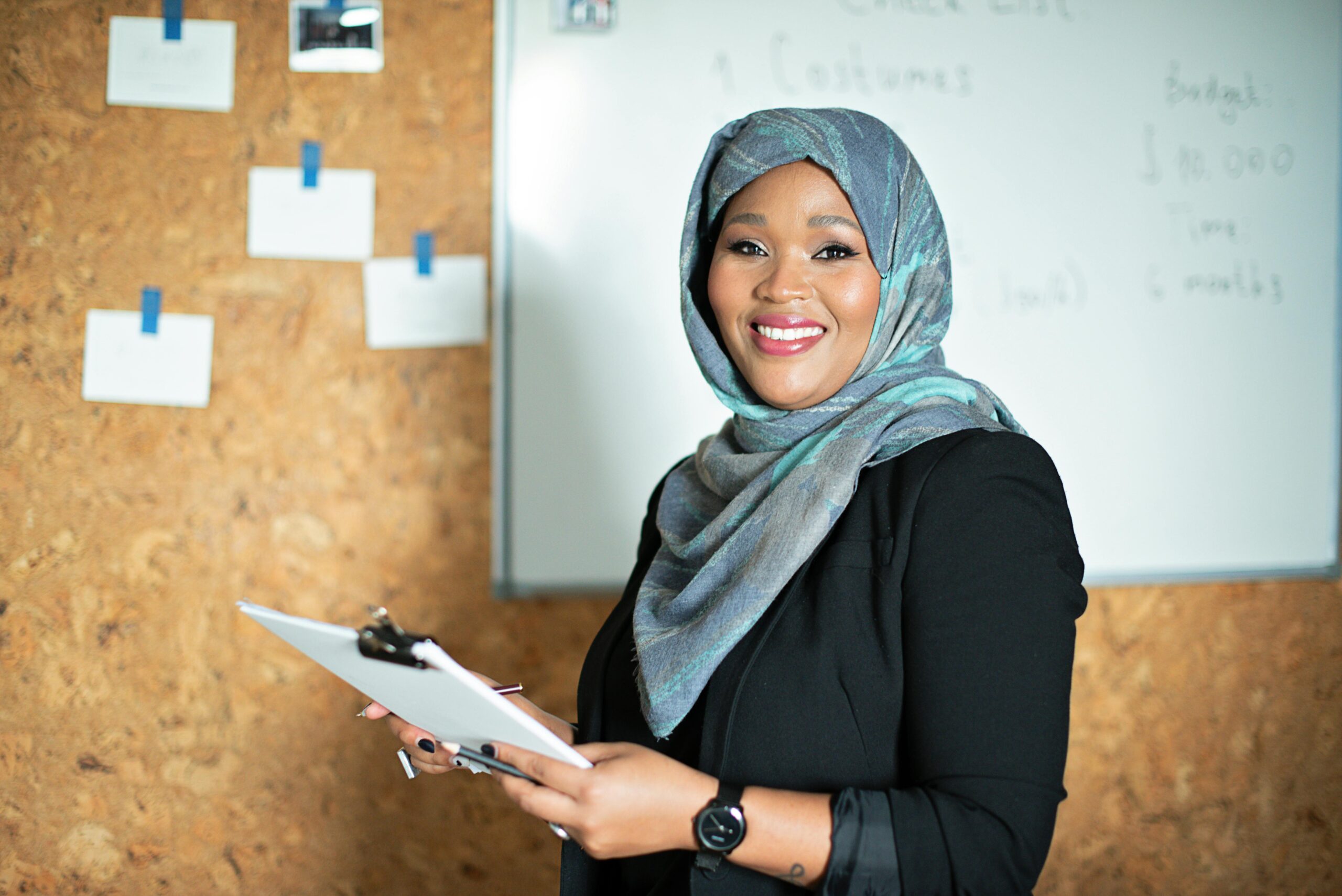 Confident businesswoman smiling while holding documents in an office setting with a whiteboard.
